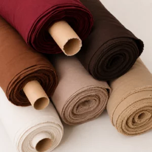 Mehrere aufgerollte Stoffbahnen aus akustischem Stoff als Meterware in den Farben Bordeaux, Dunkelbraun, Hellbraun, Sand, Beige, Hellbeige, Off-white und Weiß, dekorativ übereinander angeordnet.