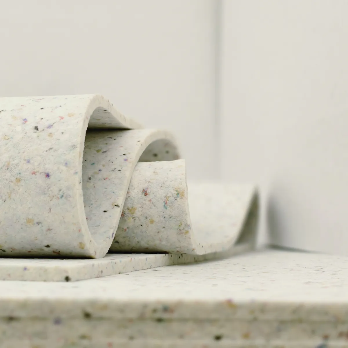 Nahaufnahme der Elitexa Teppichunterlage mit mehrfach gebogener Materialbahn auf einer flachen Unterlage, wodurch die flexible, dichte Struktur und die fein gesprenkelte Oberfläche gut sichtbar werden.