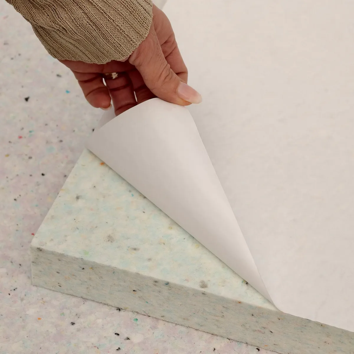 Eine Hand zieht die Schutzfolie der selbstklebenden Rückseite der naturfarbenen Elitexa Laminatboden-Unterlage ab und zeigt die dichte, faserige Materialstruktur.